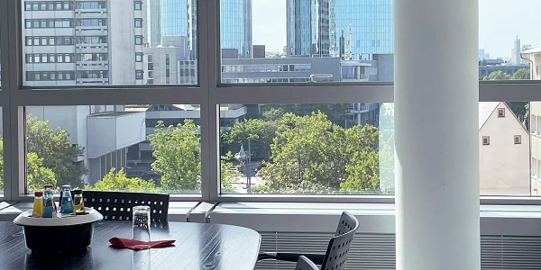 Ein Tisch mit vier Stühlen und einer Flasche Wasser steht in einem hellen Besprechungsraum, der große Fenster mit Ausblick auf moderne Gebäude und Bäume hat.