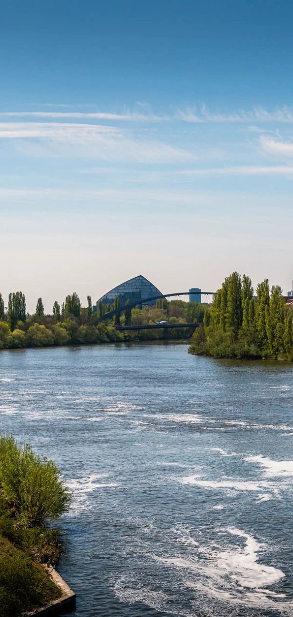 Eine breite Flusslandschaft mit ruhigem Wasser, umgeben von Bäumen und grünen Wiesen. Im Hintergrund sind moderne Gebäude und eine Brücke sichtbar, während der Himmel klar und blau ist.