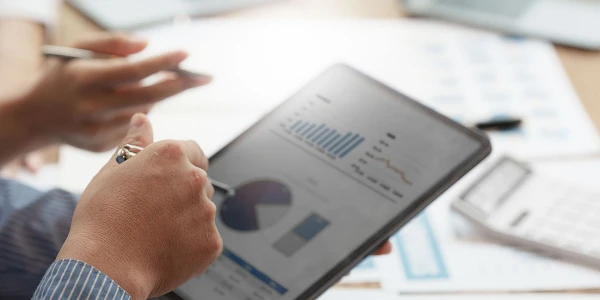 Eine Person hält ein Tablet in der Hand, auf dem Diagramme und Statistiken angezeigt werden, während im Hintergrund weitere Berichte und Grafiken auf einem Tisch liegen.