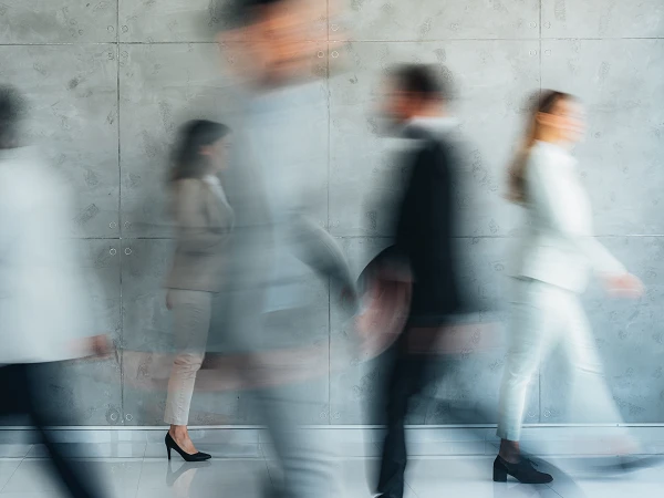 Eine Gruppe von Menschen in Geschäftsanzügen geht zügig an einer grauen Wand entlang, während sie verschwommen und in Bewegung dargestellt sind.