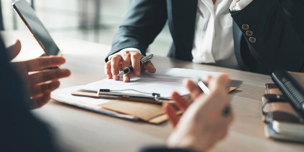 Zwei Personen sitzen an einem Tisch und diskutieren, während einer einen Stift in der Hand hält und auf Dokumente schaut. Ein Smartphone liegt auf dem Tisch, und beide konzentrieren sich auf die Unterlagen.