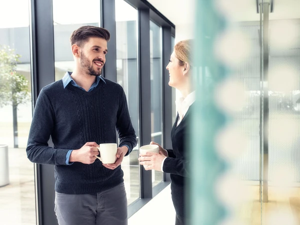 Ein Mann und eine Frau stehen in einem modernen Büroraum und unterhalten sich, während sie Tassen in der Hand halten. Große Fenster im Hintergrund lassen Tageslicht herein, und Pflanzen sind sichtbar.