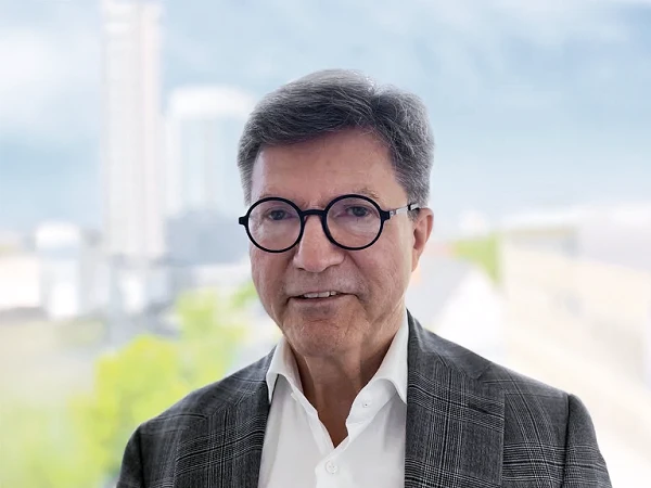 Ein älterer Mann mit Brille und grauen Haaren trägt einen grauen Blazer und ein weißes Hemd, während er in einem hellen Raum vor einer Fensteransicht steht, die eine verschwommene städtische Landschaft zeigt.