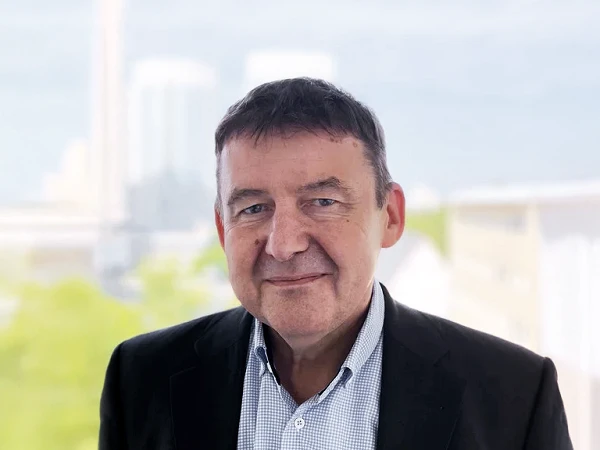 Es ist ein Mann mittleren Alters mit dunklen Haaren, der lächelt und einen schwarzen Blazer über einem karierten Hemd trägt. Im Hintergrund sind unscharfe, hellgrüne und städtische Elemente zu erkennen.