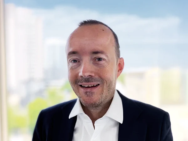 Ein Mann mit kurzen, braunen Haaren und einem freundlichen Lächeln trägt einen schwarzen Anzug und ein weißes Hemd, während er vor einem unscharfen Hintergrund aus Fenstern und städtischer Landschaft steht.