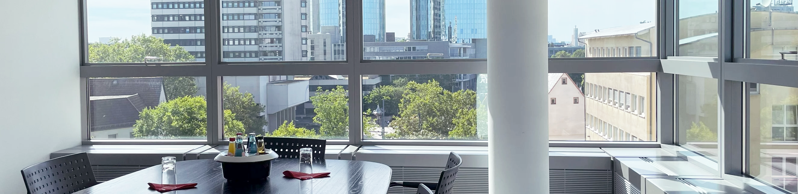 Ein Tisch mit vier Stühlen und einer Flasche Wasser steht in einem hellen Besprechungsraum, der große Fenster mit Ausblick auf moderne Gebäude und Bäume hat.