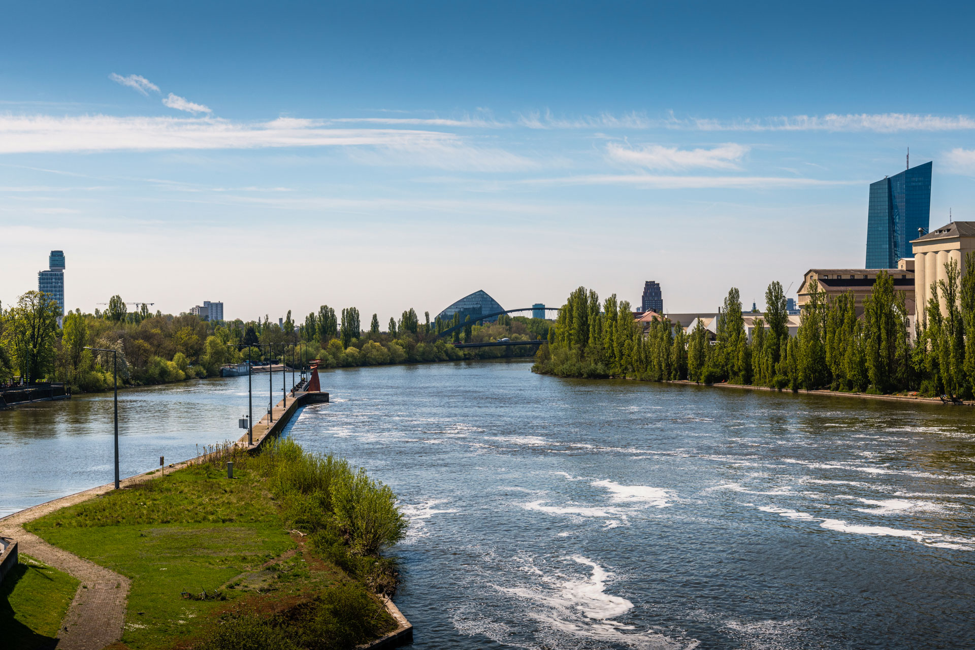 Eine breite Flusslandschaft mit ruhigem Wasser, umgeben von Bäumen und grünen Wiesen. Im Hintergrund sind moderne Gebäude und eine Brücke sichtbar, während der Himmel klar und blau ist.