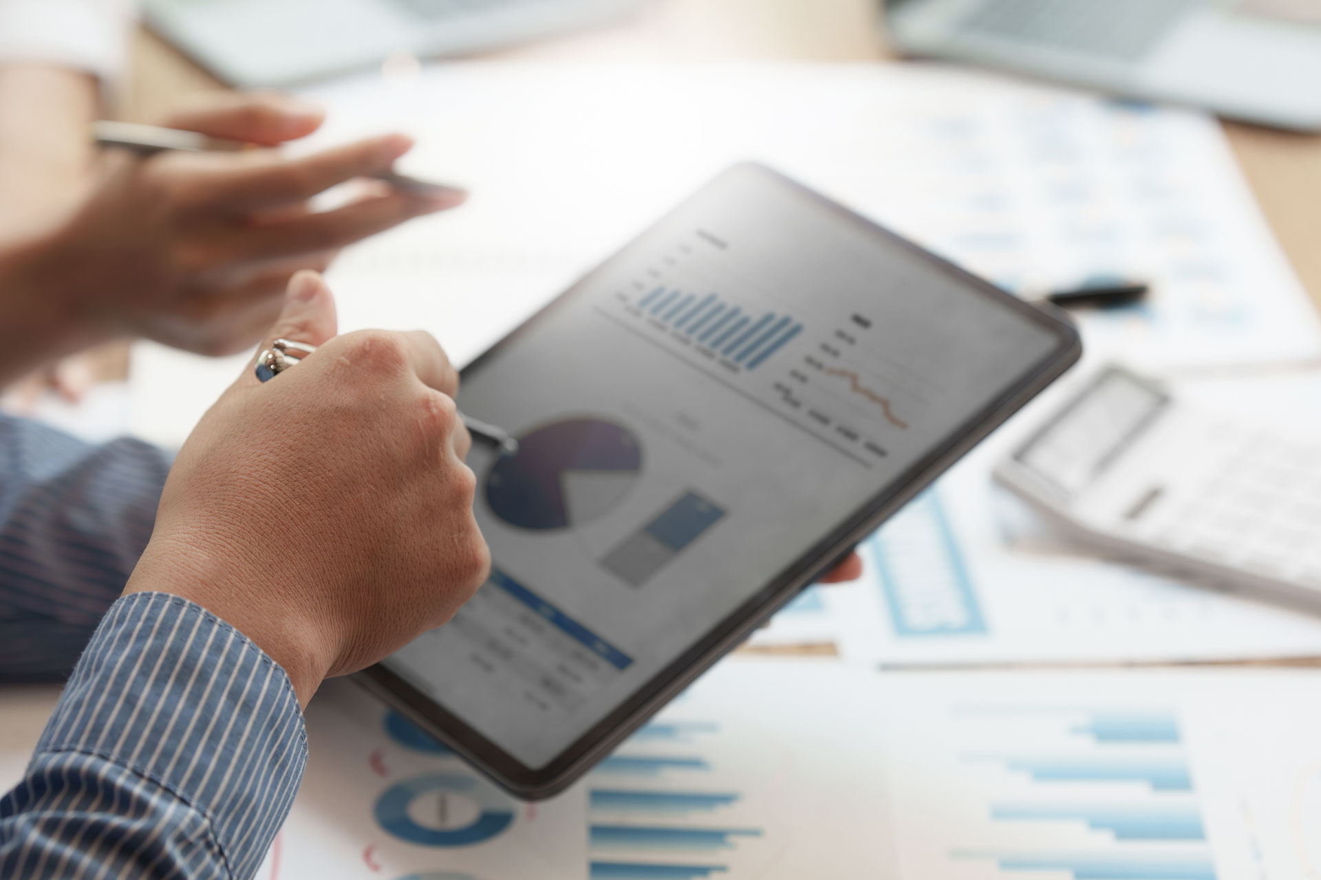 Eine Person hält ein Tablet in der Hand, auf dem Diagramme und Statistiken angezeigt werden, während im Hintergrund weitere Berichte und Grafiken auf einem Tisch liegen.