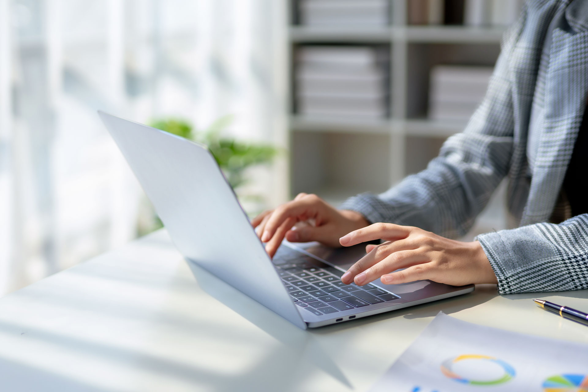 Eine Person tippt auf einem Laptop, während auf dem Tisch ein Blatt Papier mit Diagrammen und Grafiken liegt. Im Hintergrund sind Regale mit weißen Ordnern und einer Pflanze sichtbar.