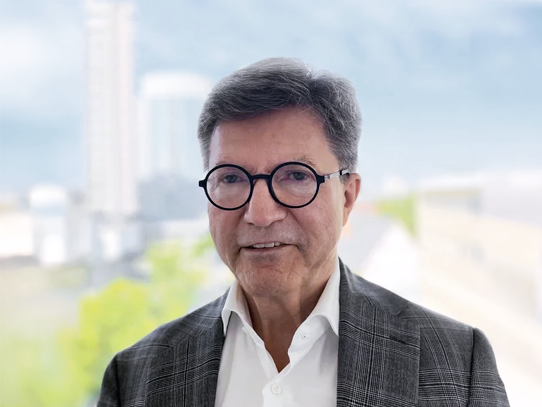Ein älterer Mann mit Brille und grauen Haaren trägt einen grauen Blazer und ein weißes Hemd, während er in einem hellen Raum vor einer Fensteransicht steht, die eine verschwommene städtische Landschaft zeigt.