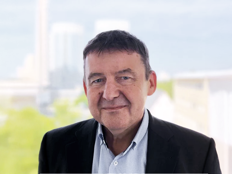 Es ist ein Mann mittleren Alters mit dunklen Haaren, der lächelt und einen schwarzen Blazer über einem karierten Hemd trägt. Im Hintergrund sind unscharfe, hellgrüne und städtische Elemente zu erkennen.